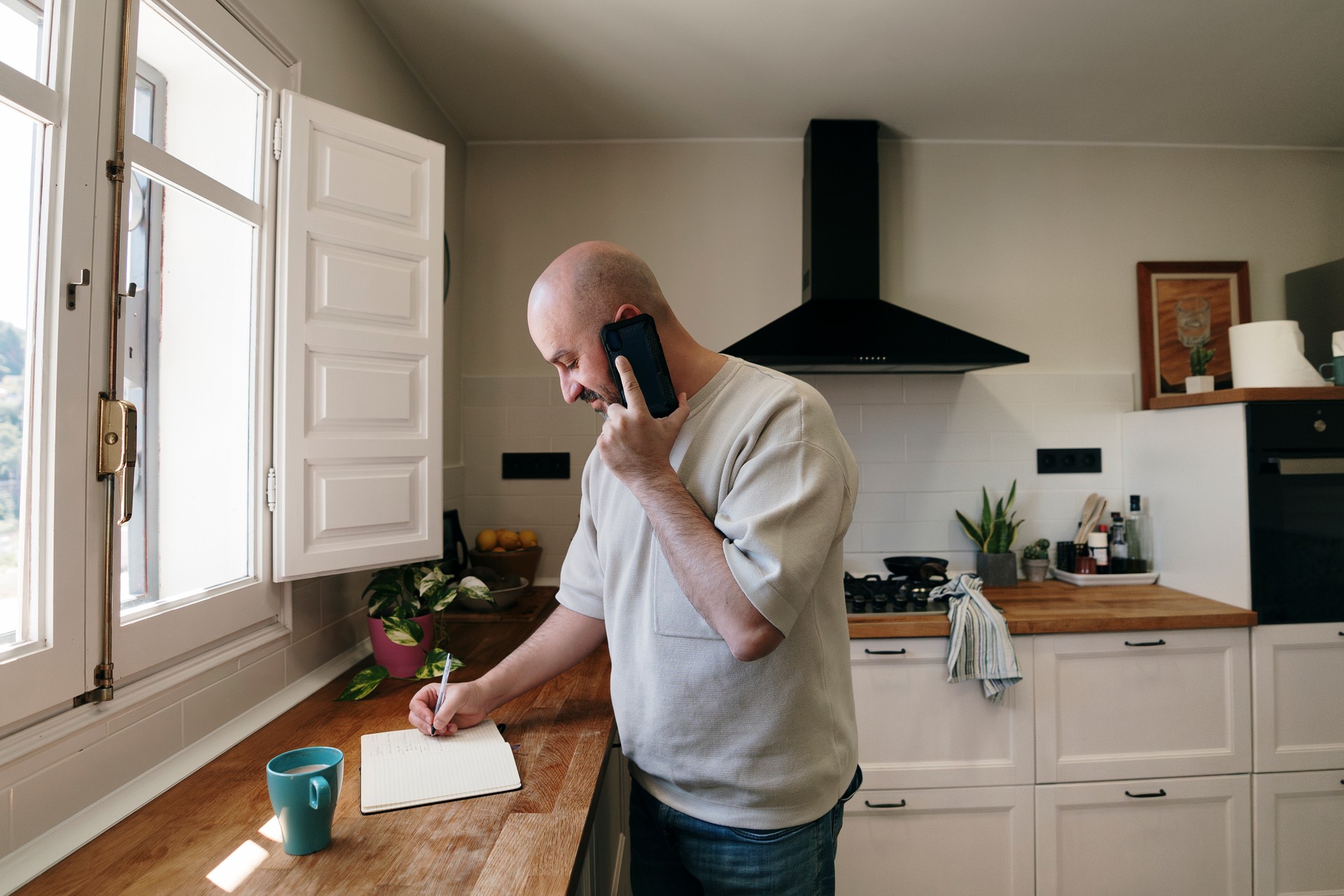 Man in kitchen writing in notebook while talking on cell phone